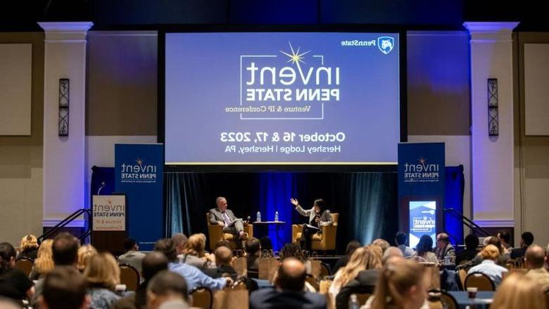 Dr. Karen Kim and Andrew Read speak sitting on a stage in front of an Invent Penn State Venture & IP Conference branded screen with a full audience in the foreground