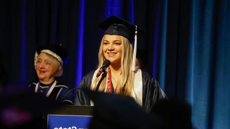 A person wearing a graduation gown stands at a lectern to speak at commencement.